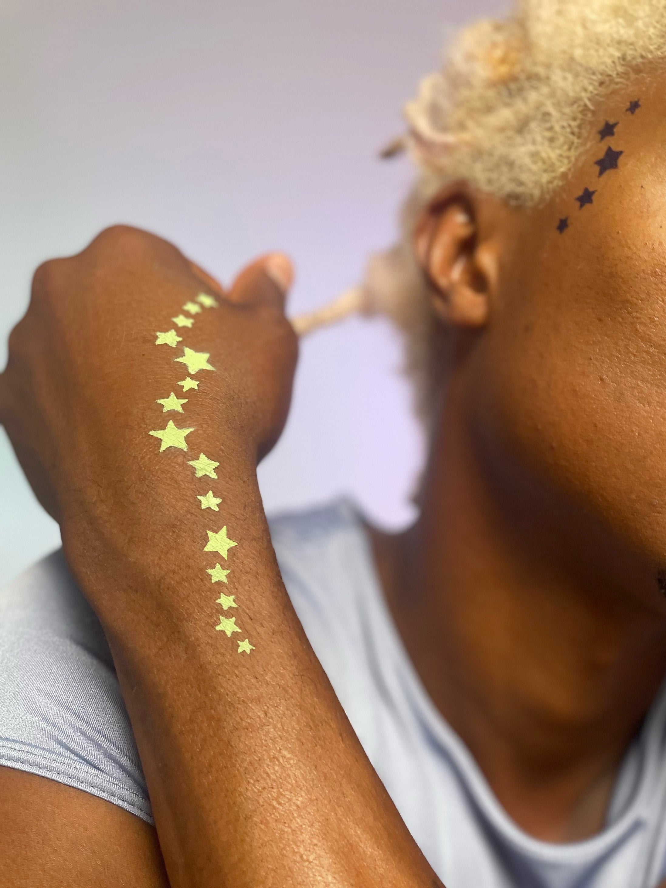 Model with dark skin wearing star pattern along inner left wrist and hand in Sagebrush and stars along temple in Corvid.